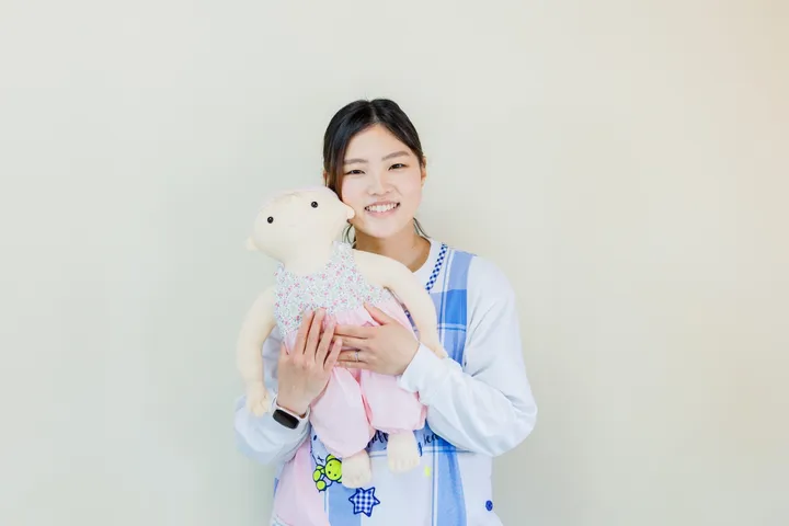 白い壁の前に立つ講師。水色と白のチェックのカーディガンを着て、ピンクの服を着せた練習用の人形を両手で胸に抱き、正面を向いて笑っている。