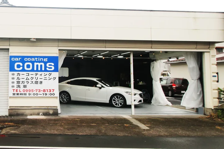 道路に面したガレージ型の店舗。左手前に「car coating COMS」の店名と施工メニューを記した看板が立ち、開口部の奥では白いセダンが照明の下に停まっている
