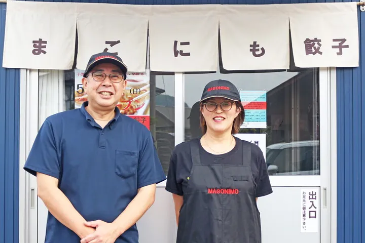 「まごにも餃子」と染め抜かれた暖簾がかかった店舗の前で、ロゴ入りの帽子をかぶった2名が並んで立っている。右側の1名はロゴ入りの黒いエプロンを着けている