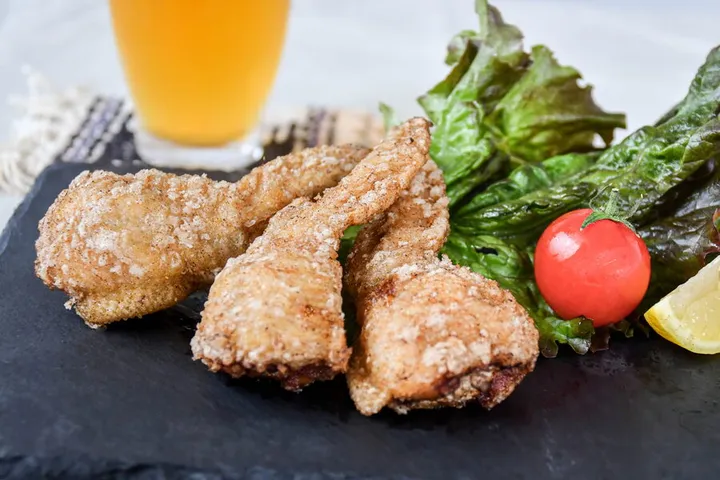 黒いスレートの皿に、粗い衣をまとって揚げられた手羽先餃子が3本並び、奥にグリーンリーフとミニトマト、レモン、グラスに入った飲み物が置かれている