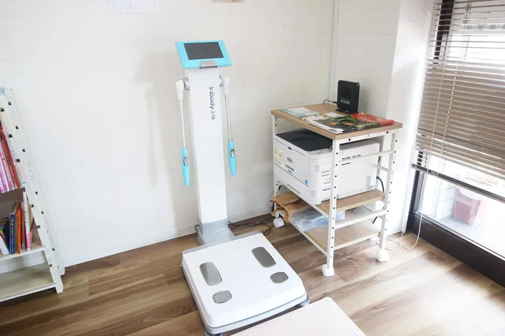 明るい部屋の壁ぎわに立つ体組成計と、その手前の床に置かれた足を乗せる測定台。右奥のワゴンにはプリンターと冊子が置かれ、左には本を並べたラックがある