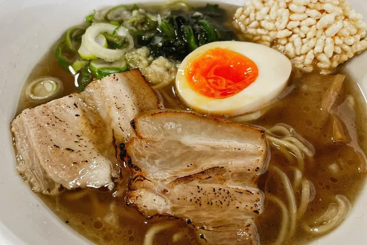 白い丼に入ったラーメンのアップ。焦げ目のついた厚切りのチャーシューが重なり、半熟の味付け卵、青ねぎ、青菜、揚げ玉のかたまりが澄んだ茶色のスープに浮かんでいる。