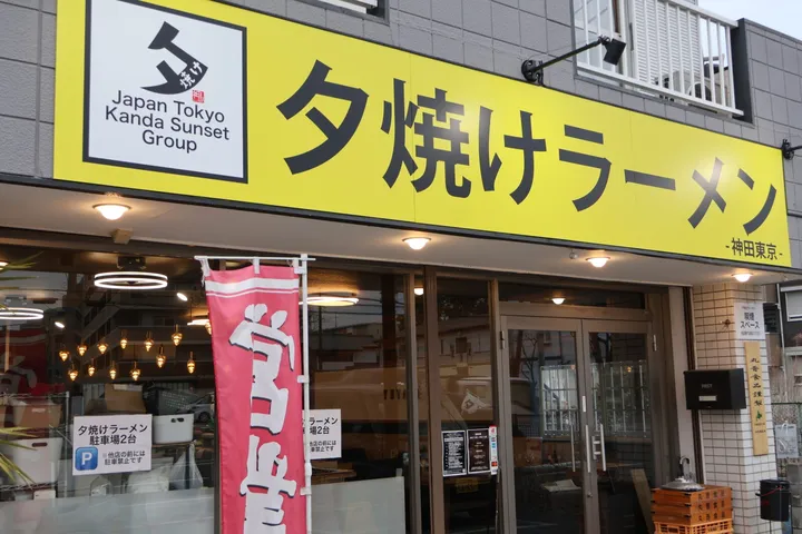 黄色い大きな看板に「夕焼けラーメン」と書かれた店舗の外観。看板の左端にグループ名のロゴが入り、ガラス張りの入口の前に赤いのぼりが立っている。右手に駐車場の案内板がある。