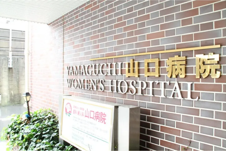 レンガ壁に「YAMAGUCHI 山口病院 WOMEN'S HOSPITAL」の金色の立体文字が掲げられた病院の入口。壁の手前に病院名を記した白い案内看板が立ち、左手に緑の植え込みがある。