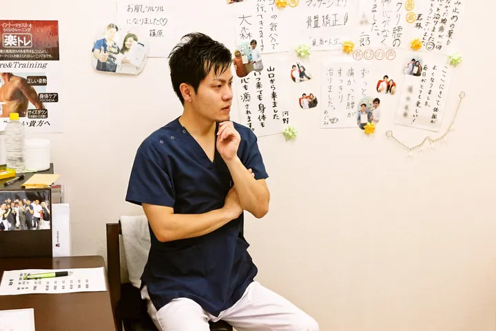 施術室で、紺色のスクラブを着た施術者が椅子に座って話している。背景の壁には来院された方からの手書きのメッセージや写真が並び、左手前には資料を置いた机がある