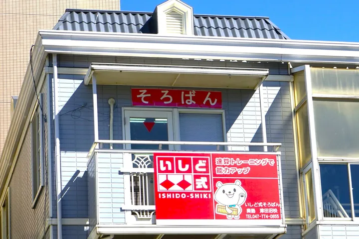 青空を背にした建物の2階部分。ベランダの上に「そろばん」と書かれた赤い看板が掲げられ、その下の柵に教室名とキャラクターが描かれた赤い看板が取り付けられている。