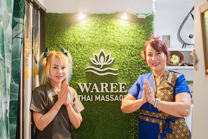 店名ロゴの入った緑の壁の前で、タイの民族衣装とユニフォーム姿のセラピスト2名が合掌して出迎えている様子