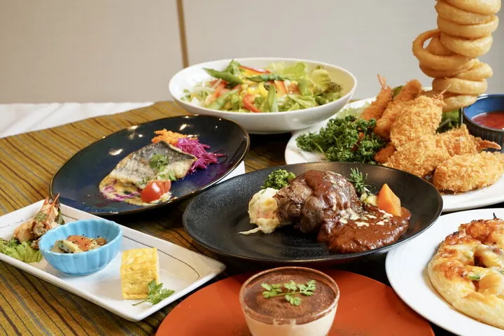 白いテーブルクロスの上に並んだ洋食の数々。手前に黒い皿のハンバーグとポテトサラダ、ティラミス、奥にサラダボウル、魚料理、エビフライとオニオンリングの盛り合わせ、ピザが置かれている