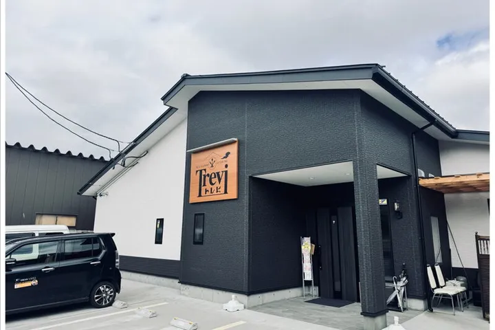 白と黒の外壁の平屋建ての店舗。入口の柱にオレンジ色の「Trevi」の看板が掛かり、手前は舗装された駐車場で、左に黒い車が停まっている