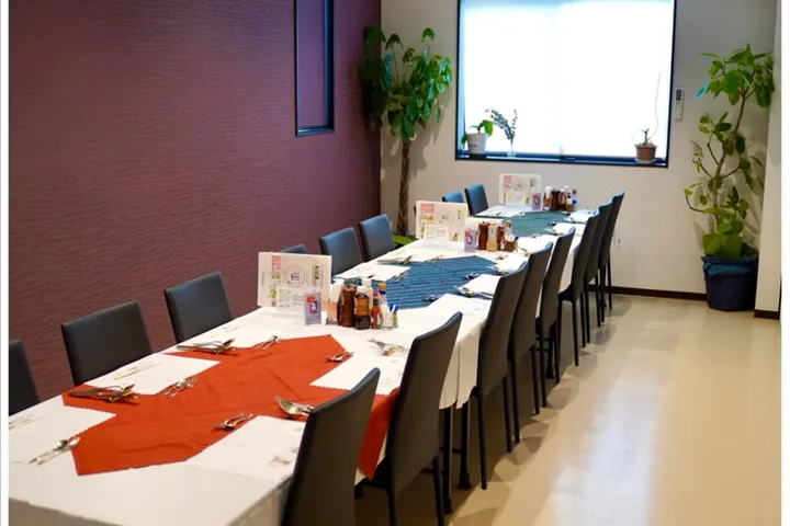 えんじ色の壁と白い床の店内。白いクロスの長いテーブルに赤と青のナプキンがセットされ、黒い椅子が両側に並ぶ。奥の窓辺に観葉植物がある