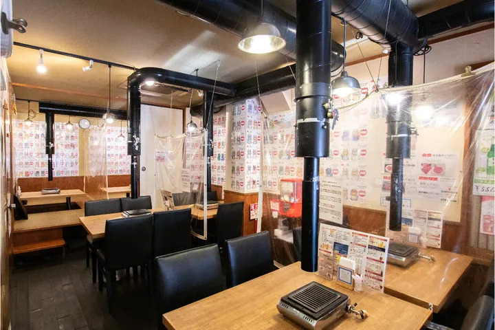 店内のテーブル席。木のテーブルごとに無煙ロースターと黒い排気ダクトが備えられ、黒い椅子が並ぶ。壁一面にメニューの札が貼られ、席のあいだに透明の間仕切りが下がっている。