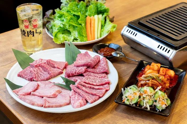 木のテーブルに並んだ焼肉の盛り合わせ。白い丸皿に赤身と霜降りの生肉が笹の葉を添えて盛られ、奥にサンチュと野菜スティックの皿、右に無煙ロースター、左に店名入りのグラス、右手前にキムチとナムルの小鉢が置かれている。